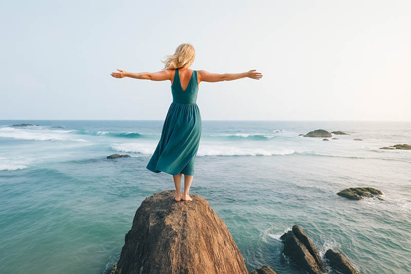 Woman standing on rock by the ocean with arms outstretched - representing freedom and healing through hypnotherapy